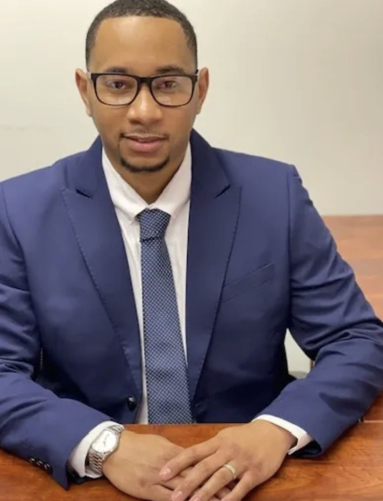 Headshot a Black man in a blue suit and tie with glasses on, hands crossed, sitting at a wooden table.