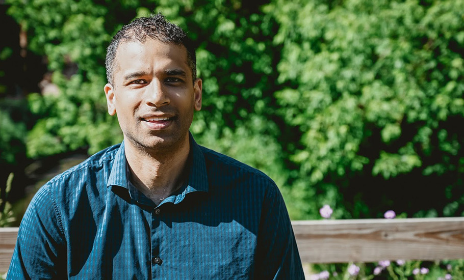 South asian man in a blue shirt smiling against a background of greenery.