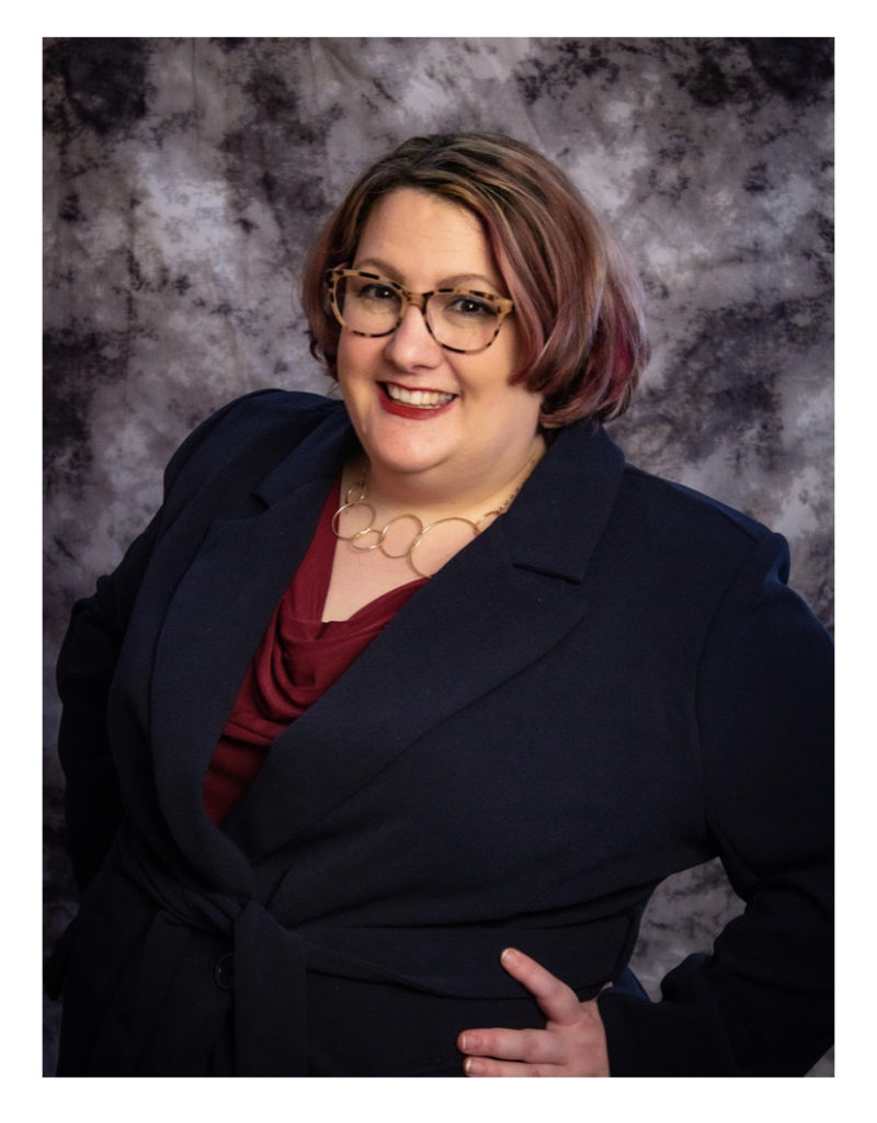 Professional photo of a white woman with short brown hair, smiling in front of a dappled grey and white background, wearing a navy suit with a red shirt.