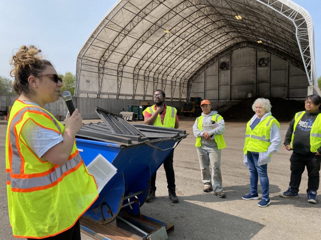 Maria Bianchetti stands with a microphone next to a residential compost drop off bucket.