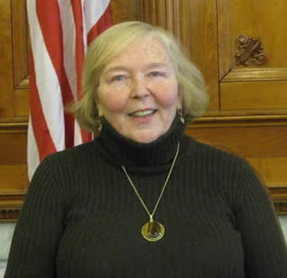 White woman in black turtleneck against a wood-panelled background with a small amount of American flag showing.