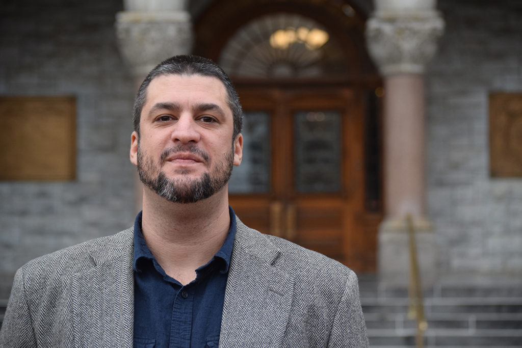 Headshot of a white man with a beard and short hair wearing a grey/tan blazer against a backdrop of an older building with heavy wooden doors.