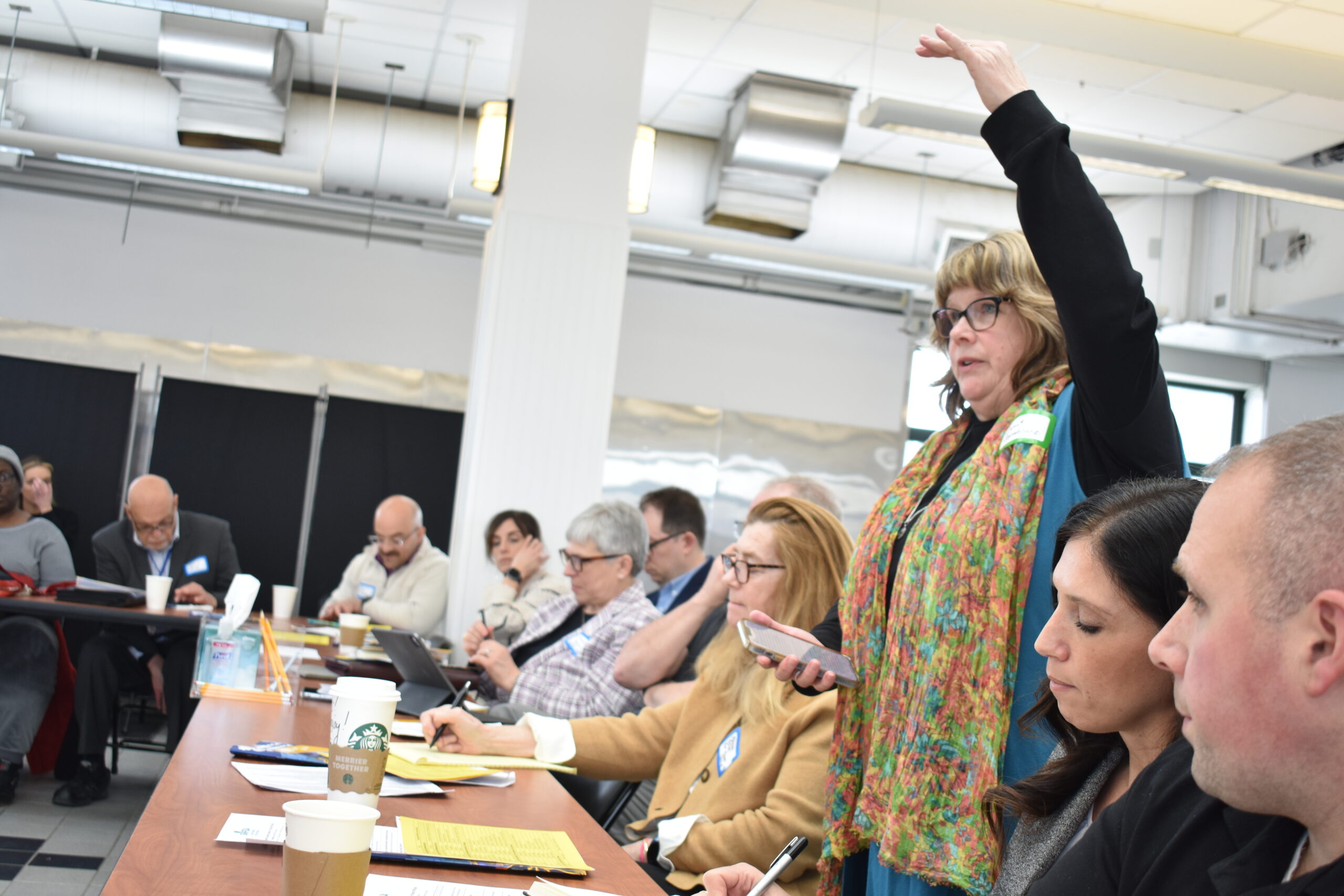 White woman stands up, gestures with hand to indicate scale, in a room full of seating people