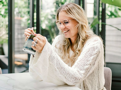 Marieke Rijksen (Whispering Bold) sitting at a marble table, smiling while stirring a drink in a ceramic mug. She is wearing glasses and a loose-knit cream sweater. Large glass doors and lush green plants create a bright, airy background.