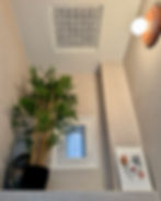 Indoor scene with a bamboo plant, abstract art of hands and face, small window, decorative ceiling vent, and wall light. Calm, minimalist vibe.