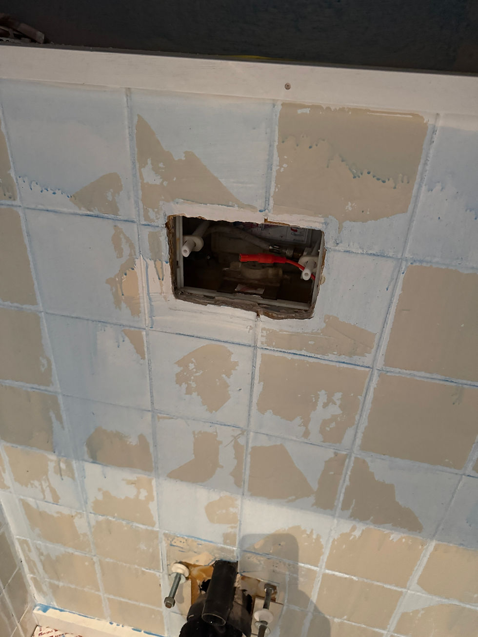 A wall with partially stripped pale tiles shows an exposed plumbing pipe and fittings in a square opening, suggesting renovation work.