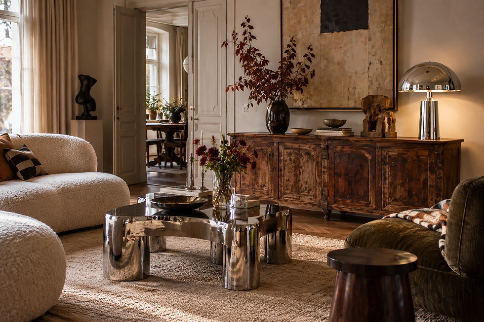 Cozy living room with cream sofas, chrome coffee table, rustic sideboard, abstract art, and warm sunlight.