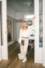 Smiling woman in a cozy room, wearing a white sweater and beige pants, leans on a door frame holding a blue folder. Elegant decor in background.