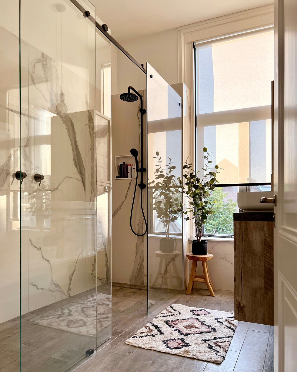 Modern bathroom with glass shower, marble walls, wood floor, and soft rug. Potted plant on stool by window adds a fresh touch.