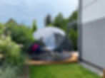 Clear geodesic dome with chairs and tables inside on a wooden deck, surrounded by greenery. Part of a house wall is visible.