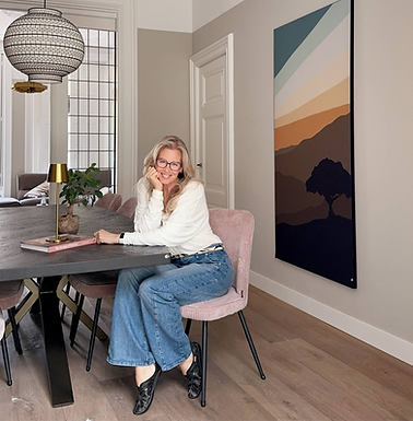 A woman with long blonde hair and glasses sits at a modern dining table, smiling warmly. She wears a white sweater, blue jeans, and black shoes. The dining room features soft neutral tones, with a black dining table, pink upholstered chairs, and a gold-accented table lamp. A large, abstract landscape artwork hangs on the wall, and an elegant pendant light with a woven design hangs from the ceiling. The space has classic architectural details, including decorative ceiling mouldings and tall doors. In the background, a living area with a sofa and a glass-paneled door is visible.