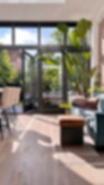 Stylish living room with large glass doors opening to a garden. Features a green plant, blue sofa with patterned cushions, and bar stools.