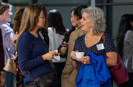 Two professional women networking at a business event, engaging in conversation while holding coffee and refreshments.