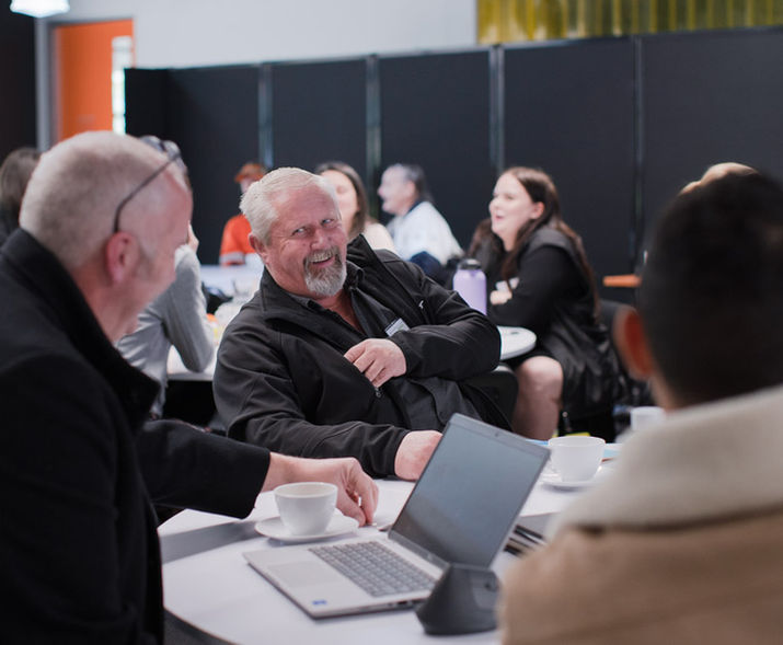 Group of professionals networking at a business event, smiling and engaging in conversation around tables with laptops and coffee cups.