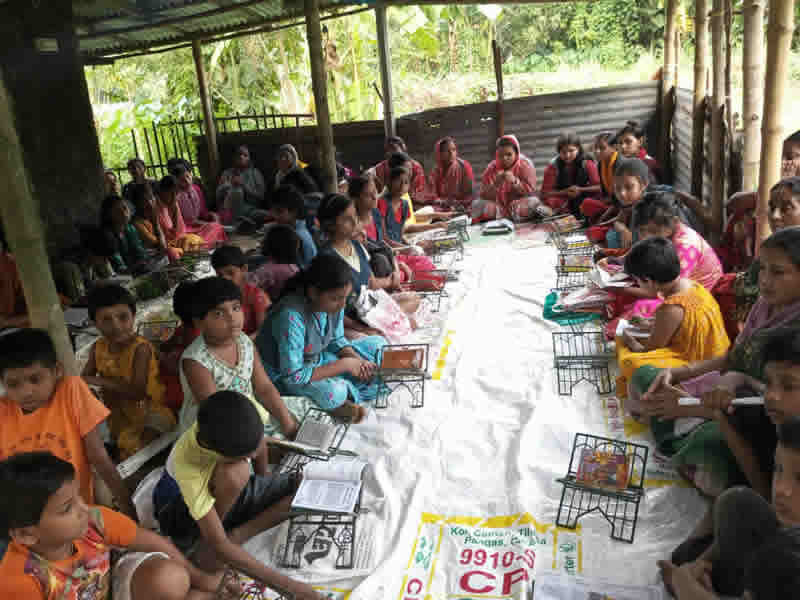 Children and adults gathered in an open-air structure with a bamboo or wooden frame roof and natural surroundings visible in the background. They are seated on the floor in rows on either side of a long white cloth or sheet laid down the center. Various educational materials including small wire frame models (possibly geometric shapes or learning tools), papers, and teaching materials are placed along the cloth. The participants are wearing colorful clothing in shades of pink, orange, blue, yellow, and other bright colors. The setting appears to be a community learning space or informal classroom in a rural area.