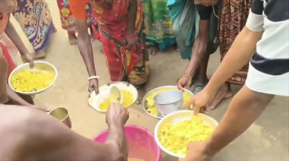 "People serve and hold plates of khichdi from bowls on the ground, surrounded by a crowd of women waiting to receive food."