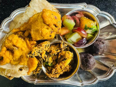 A decorative metal serving tray displays an assortment of Indian vegetarian dishes. The tray contains golden-yellow cauliflower curry, flatbread, a colorful mixed vegetable salad with cucumbers and tomatoes, a spiced lentil or chickpea dish garnished with herbs, and dark round sweets or dessert balls. The food is arranged in small metal bowls on an ornate silver-toned serving platter against a dark surface.