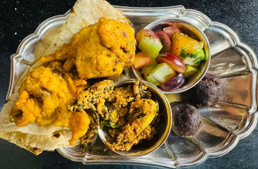 A decorative metal serving tray displays an assortment of Indian vegetarian dishes. The tray contains golden-yellow cauliflower curry, flatbread, a colorful mixed vegetable salad with cucumbers and tomatoes, a spiced lentil or chickpea dish garnished with herbs, and dark round sweets or dessert balls. The food is arranged in small metal bowls on an ornate silver-toned serving platter against a dark surface.