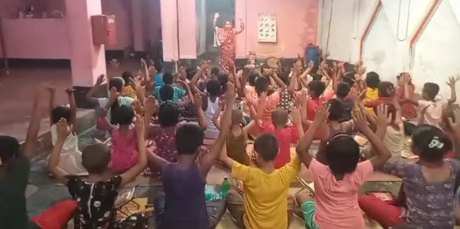 A large group of children sitting on the floor in rows with their arms raised up in the air during what appears to be a group activity or exercise session. An instructor in patterned clothing stands elevated at the front of the room. The setting appears to be an indoor community space or classroom with pink and white walls, and the children are wearing colorful casual clothing in shades of pink, yellow, blue, and other bright colors.