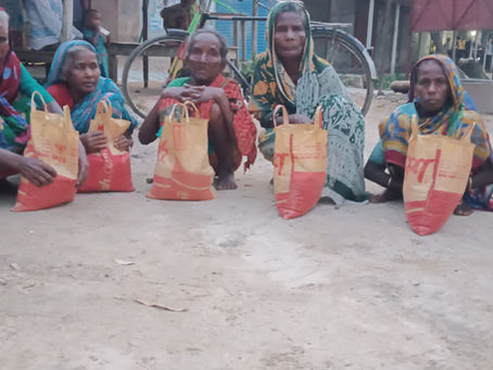 A group of elderly women sitting on the ground outdoors, each with a large orange and yellow bag of rice in front of them. The women are dressed in colorful sarees and head coverings, waiting during what appears to be a food aid or charitable distribution event in a rural South Asian community.
