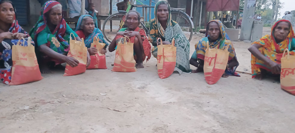 A group of elderly women sitting on the ground outdoors, each with a large orange and yellow bag of rice in front of them. The women are dressed in colorful sarees and head coverings, waiting during what appears to be a food aid or charitable distribution event in a rural South Asian community.