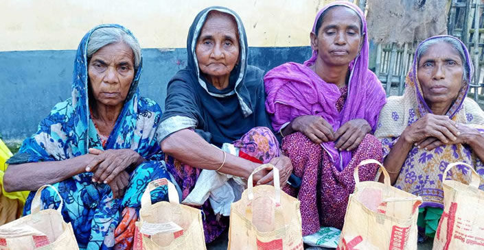 Four elderly women sitting together, each wearing colorful head coverings and traditional clothing. From left to right: a woman in a blue patterned sari and head covering, a woman in a dark blue/navy head covering, a woman in a bright purple/magenta sari and head covering, and a woman in a light-colored sari with floral patterns. In front of them are several white cloth or paper bags with red text or logos, appearing to be distribution bags or relief supplies. The women are seated against what appears to be a blue and white wall, suggesting this may be part of a community aid or distribution program.