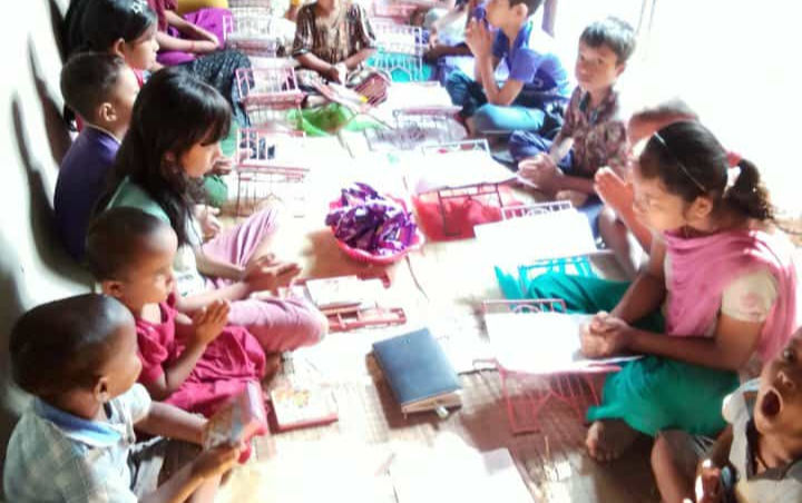 A group of children and several young women sit closely together on woven mats in a narrow open-air classroom, studying from notebooks and books. The students appear engaged in lessons while seated in rows along the walls. Sunlight streams in from the open side of the structure, creating bright patches across the floor. Some children look toward the camera while others focus on their work. Bengali text and a date appear in the upper-left corner of the image.