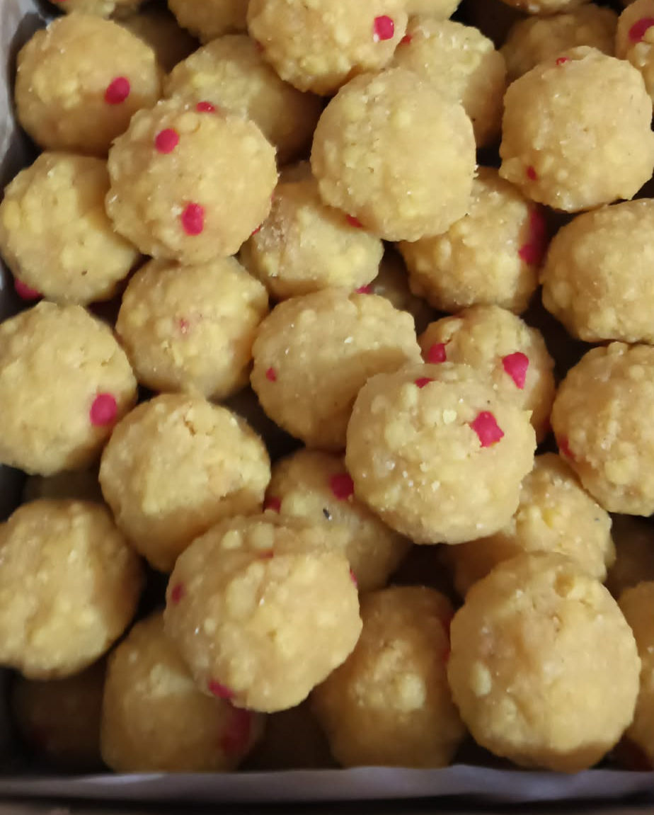 A box of golden yellow besan laddus dotted with pink sugar sprinkles, freshly made and piled together.