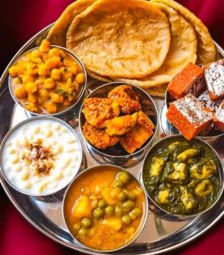 A traditional Indian thali served on a stainless steel plate with multiple small bowls containing various dishes. The meal includes flatbread (likely chapati or roti), white rice with what appears to be ghee or clarified butter, yellow dal or curry, orange-colored vegetables (possibly cauliflower or potatoes), green leafy vegetables (likely palak or spinach-based dish), a pea and potato curry, orange-colored sweets or desserts (possibly carrot halwa or similar), and what appears to be yogurt or raita. The colorful array of dishes represents a complete, balanced Indian meal with various textures, flavors, and nutritional components arranged in the traditional thali style.