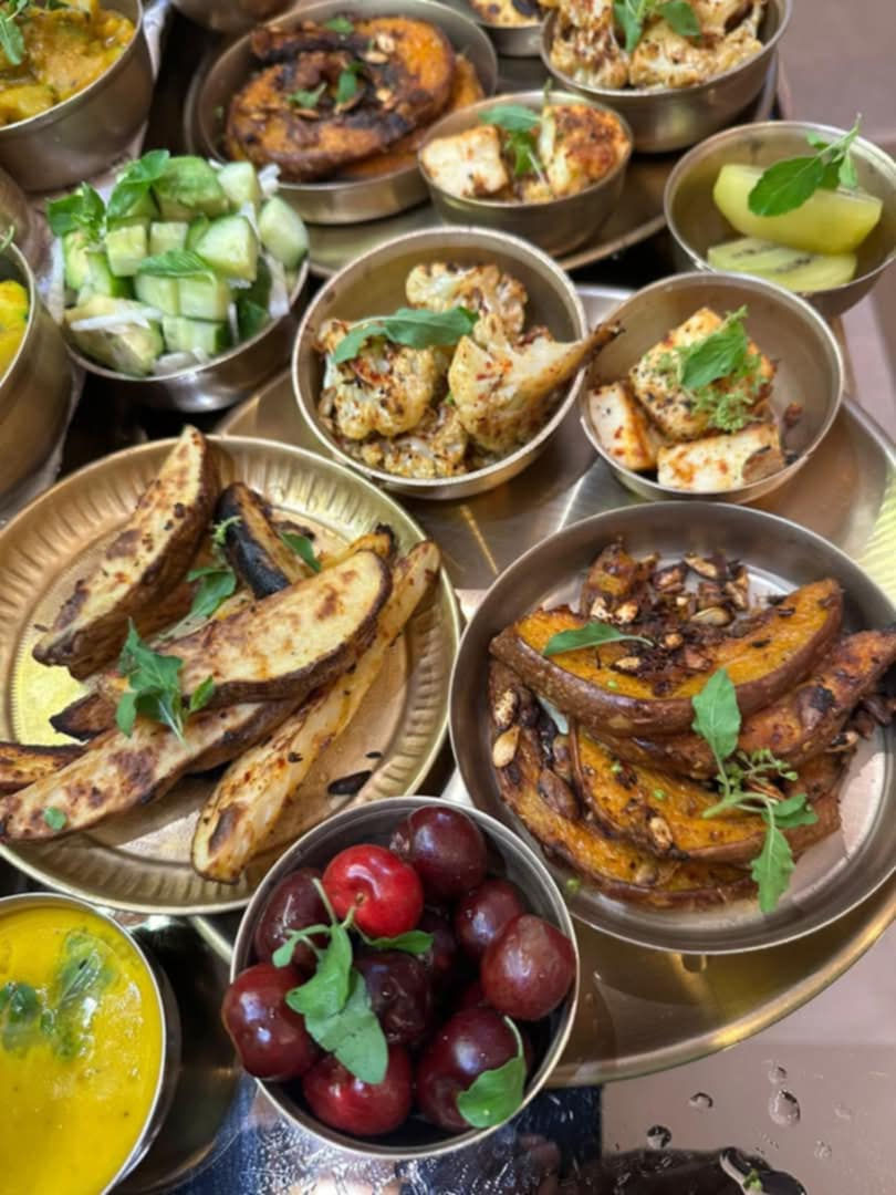 An overhead view of a plant-based meal arranged on a large brass or golden-colored tray. The meal includes multiple small bowls and plates containing: grilled eggplant slices with nuts and herbs, stuffed vegetables (possibly stuffed zucchini or grape leaves), fresh red cherries, sliced cucumbers, roasted potato wedges, what appears to be kibbeh or stuffed bulgur balls, pickled vegetables, and various other traditional dishes. Most items are garnished with fresh green herbs, likely parsley or mint. The dishes are served in small metal bowls and plates, creating a diverse array of colors and textures typical of Middle Eastern cuisine. Yellow sauces or dips are visible in some of the bowls, and the overall presentation suggests a traditional family-style or mezze-style meal.