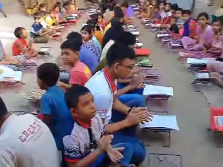 A large group of children and adults are seated on the ground in rows for what appears to be an outdoor educational or community event. Each person has books or learning materials placed in front of them on small individual mats or surfaces. The participants include children of various ages wearing colorful clothing - blue, red, pink, and other bright colors. Some appear to be wearing school uniforms or matching outfits with red accents. The setting looks like it's taking place on a concrete or paved outdoor area, possibly a school courtyard or community center. The organized arrangement and presence of educational materials suggests this could be a mass reading program, literacy initiative, or educational ceremony taking place in what appears to be a developing region.