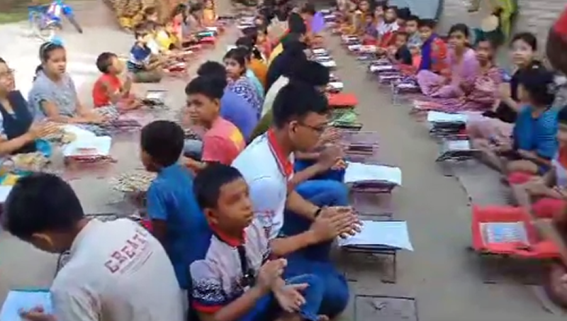 A large group of children and adults are seated on the ground in rows for what appears to be an outdoor educational or community event. Each person has books or learning materials placed in front of them on small individual mats or surfaces. The participants include children of various ages wearing colorful clothing - blue, red, pink, and other bright colors. Some appear to be wearing school uniforms or matching outfits with red accents. The setting looks like it's taking place on a concrete or paved outdoor area, possibly a school courtyard or community center. The organized arrangement and presence of educational materials suggests this could be a mass reading program, literacy initiative, or educational ceremony taking place in what appears to be a developing region.