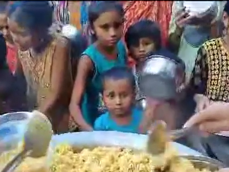 A group of children gathered around a large metal tray filled with yellow rice and curry, waiting to be served food. Several children in colorful clothing stand in line while hands reach toward the tray to distribute the meal. The scene appears to be a community food distribution event serving children in need.