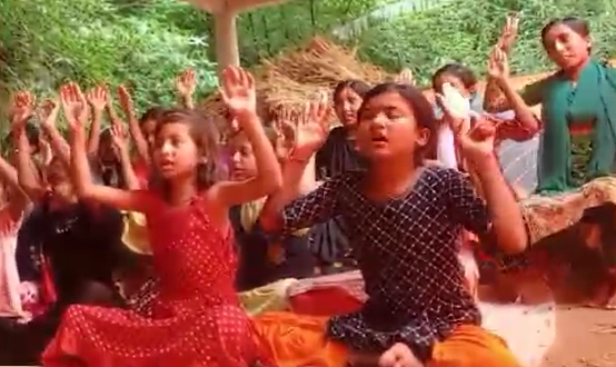 "Children sit outdoors under a shelter, raising their hands and singing together during a class."