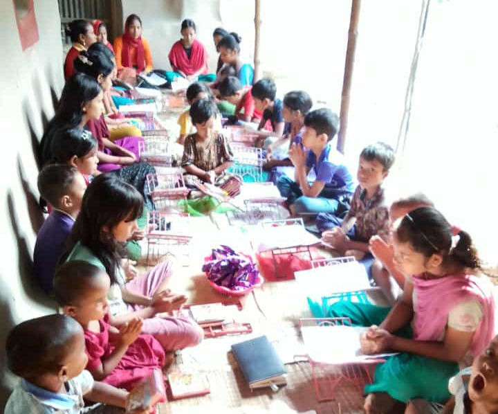 A group of children and several adults sit cross-legged on a floor covered in mats inside a simple, sunlit building. They are arranged in two rows facing each other, many with their hands together in a prayer-like gesture. Small metal book stands are placed in front of each student.