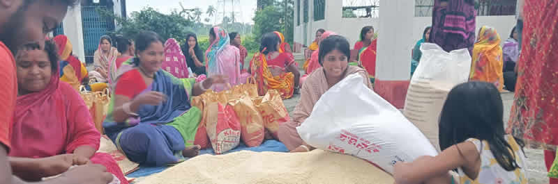 A group of women and some men sitting on the ground in what appears to be a community gathering or distribution event. Many of the women are wearing colorful traditional clothing including saris and head coverings in shades of pink, red, yellow, and white. There are white bags or packages visible that appear to contain distributed items, with some text visible on the bags. The setting looks like it's taking place outdoors or in a covered area with greenery visible in the background. The people appear to be participating in some kind of community program or aid distribution.