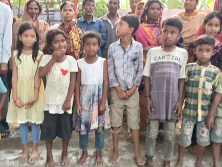 A group of children and adults in colorful clothing gather outside a rural community building. Many of the barefoot children stand together in rows, while adults stand behind them.
