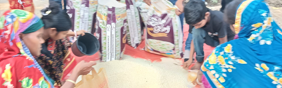 Volunteers distribute uncooked rice to villagers by scooping it from a large pile into reusable bags. Women and children gather around the rice while large sacks of rice stand in the background, illustrating a community food relief effort.