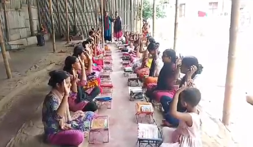 A group of children sitting in rows on the floor of what appears to be a covered outdoor area or veranda, each with small metal lunch boxes or food containers placed in front of them. The children are wearing colorful clothing and appear to be participating in a communal meal or food distribution program. The setting has concrete flooring and pillars, with a corrugated metal or bamboo wall structure visible in the background. Natural light filters in from the open sides of the covered area. The organized arrangement suggests this is part of a structured feeding program or community meal initiative for children