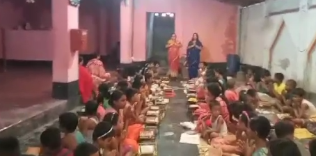 A large group of children sitting in rows on the floor of what appears to be a community hall or large indoor space, sharing a communal meal. The children are seated along long lines with metal plates or bowls in front of them, suggesting an organized meal service. Two adults in traditional clothing - one wearing an orange/saffron colored garment and another in blue - stand supervising the meal. The room has pink and white walls with some windows visible in the background. The setting appears to be in a school, community center, or charitable organization where meals are being provided to children.