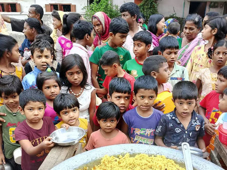 A large group of children and adults gathered around a table for a community meal distribution. In the foreground, a large metal pan contains yellow rice or similar prepared food. Children of various ages, wearing colorful clothing including green, purple, pink, and other bright colors, are holding plates and containers as they wait to be served. Adult women in traditional dress, including some wearing headscarves, supervise the meal service. The setting appears to be outdoors in a courtyard or community space with white buildings visible in the background. This appears to be a community feeding program or charitable meal distribution event.
