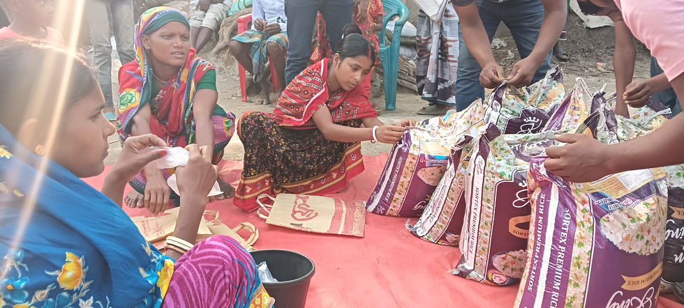 Women and a young girl sit on a red mat outdoors, handling large bags of Sortex Premium Rice during a community food relief distribution, with additional bags and volunteers visible in the background
