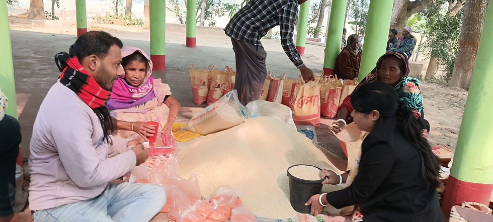 Volunteers and community members sit on the ground under a covered outdoor pavilion with green pillars, scooping rice from a large pile into bags and plastic packaging for distribution. Several filled burlap sacks are lined up in the background.