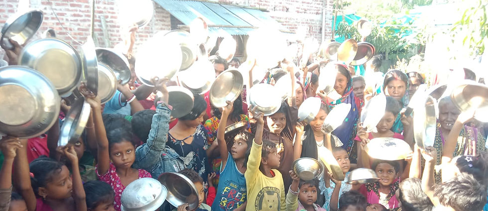 "A crowd of children and women hold up empty metal plates and bowls above their heads around a large pot of khichdi."