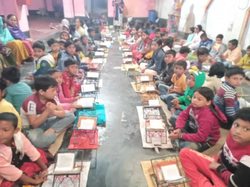 Children seated in parallel rows on the floor of a large indoor space, with low wooden desks or book stands placed in front of each child. The students are wearing colorful winter clothing including sweaters and jackets in shades of pink, red, yellow, green, and blue. Books or reading materials are positioned on the stands before them. Adult supervisors can be seen in the background. The setting appears to be a community learning center or educational program with pink-toned walls.