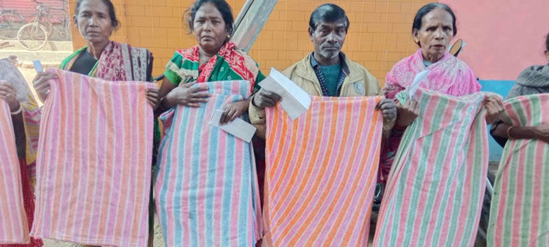 Four elderly people, three women and one man, standing in a row and holding up colorful striped fabric or textile pieces. The fabrics display various striped patterns in colors including pink, orange, green, and other bright hues. The women are wearing traditional Indian clothing - saris and blouses in colors like pink, green, and patterned fabrics. The man is wearing a light-colored shirt or jacket. They appear to be displaying their textile work or products, possibly as part of a craft demonstration or community program. The setting appears to be outdoors with buildings visible in the background.
