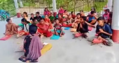 A group of children sitting on the floor in a circle outdoors, appearing to participate in a group activity or class session. The children are wearing colorful clothing in shades of pink, blue, and other bright colors. They are seated on what appears to be a concrete surface with a white building wall and metal fencing visible in the background. An adult or instructor appears to be seated with the group, facilitating the activity.