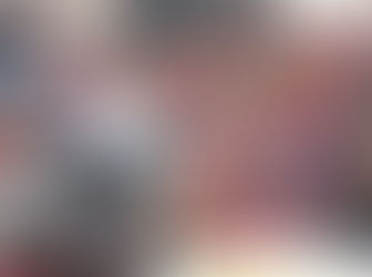 A large group of children and community members gathered outdoors in front of a brick building, standing around a large metal cooking pot containing kichri. Many children are holding empty metal plates and bowls, waiting to be served. The group includes people of various ages wearing colorful winter clothing including hooded sweatshirts and jackets in shades of pink, red, orange, and blue. The scene appears to be a community meal distribution event