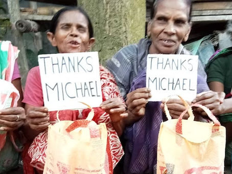 Four women sitting together outdoors, each holding handwritten "THANKS MICHAEL" signs. The women appear to be of varying ages, wearing colorful traditional clothing in shades of pink, red, purple, and green. They are smiling at the camera, and the setting appears to be in a rural or village environment with natural materials and structures visible in the background.