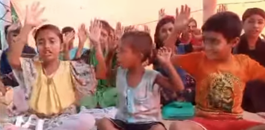 "Children sit together and raise their hands joyfully while chanting during a Bhagavad Gita class in Bangladesh, with books and supplies visible in front of them." 